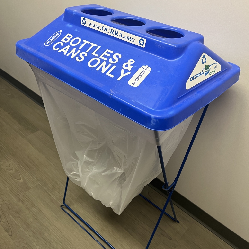 A container with a clear pastic bag on the bottom is capped by a hard plastic top with holes that give people room to drop cans and bottles inside the bag below. The top says Bottles and Cans only and has images of a bottle and can.